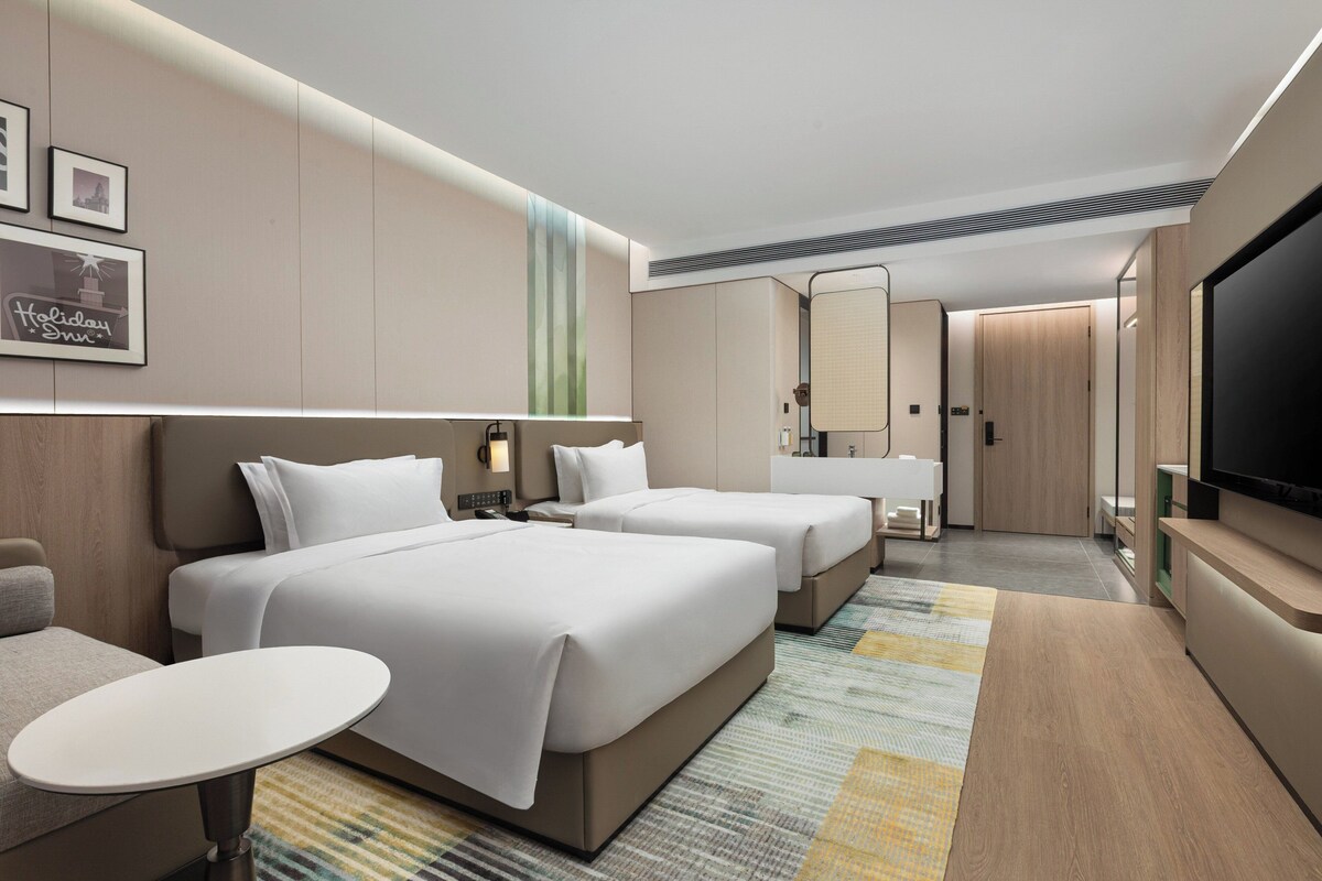 A hotel room with two beds, a small table, a TV, and a bathroom with a mirror and sink.