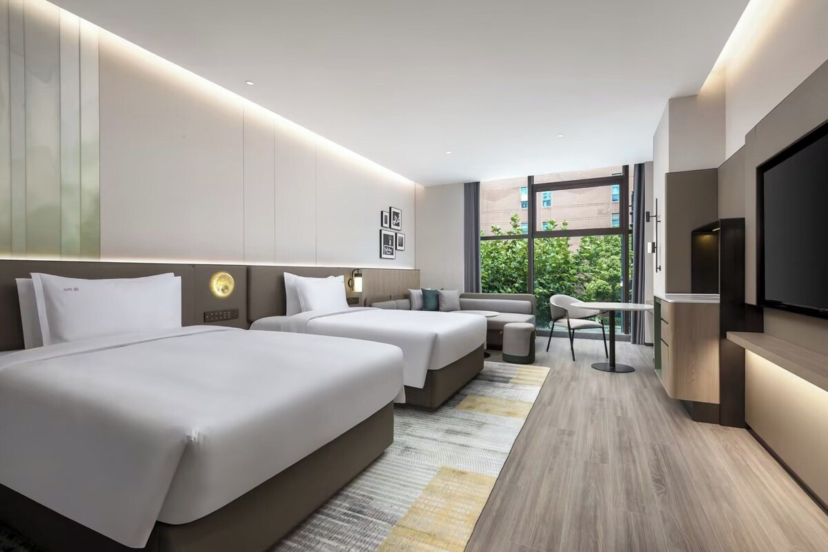 A modern hotel room with two beds, a sofa, a TV, and a view of greenery outside.