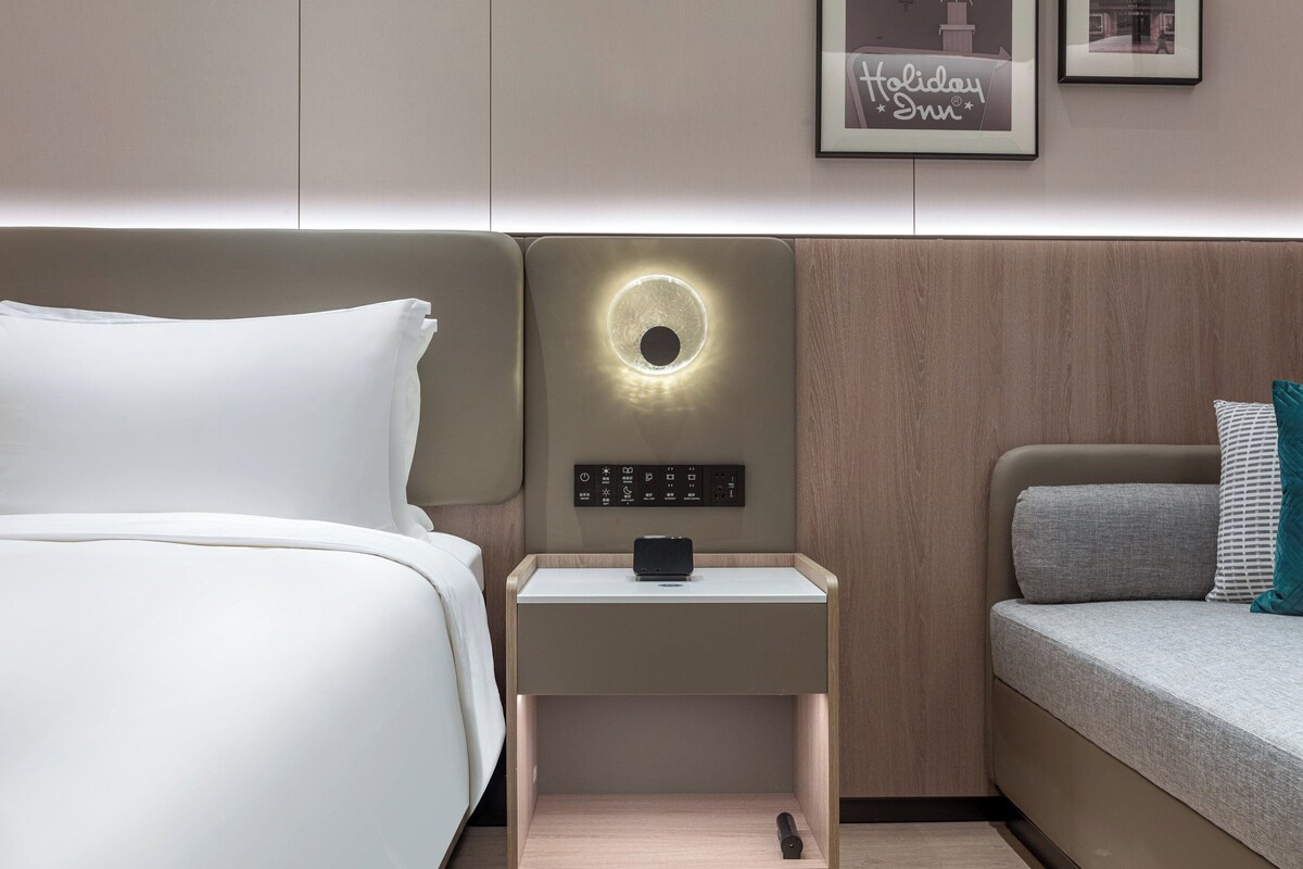 A modern hotel room with a bed, a bedside table with a built-in lamp, a control panel, and a framed picture on the wall.