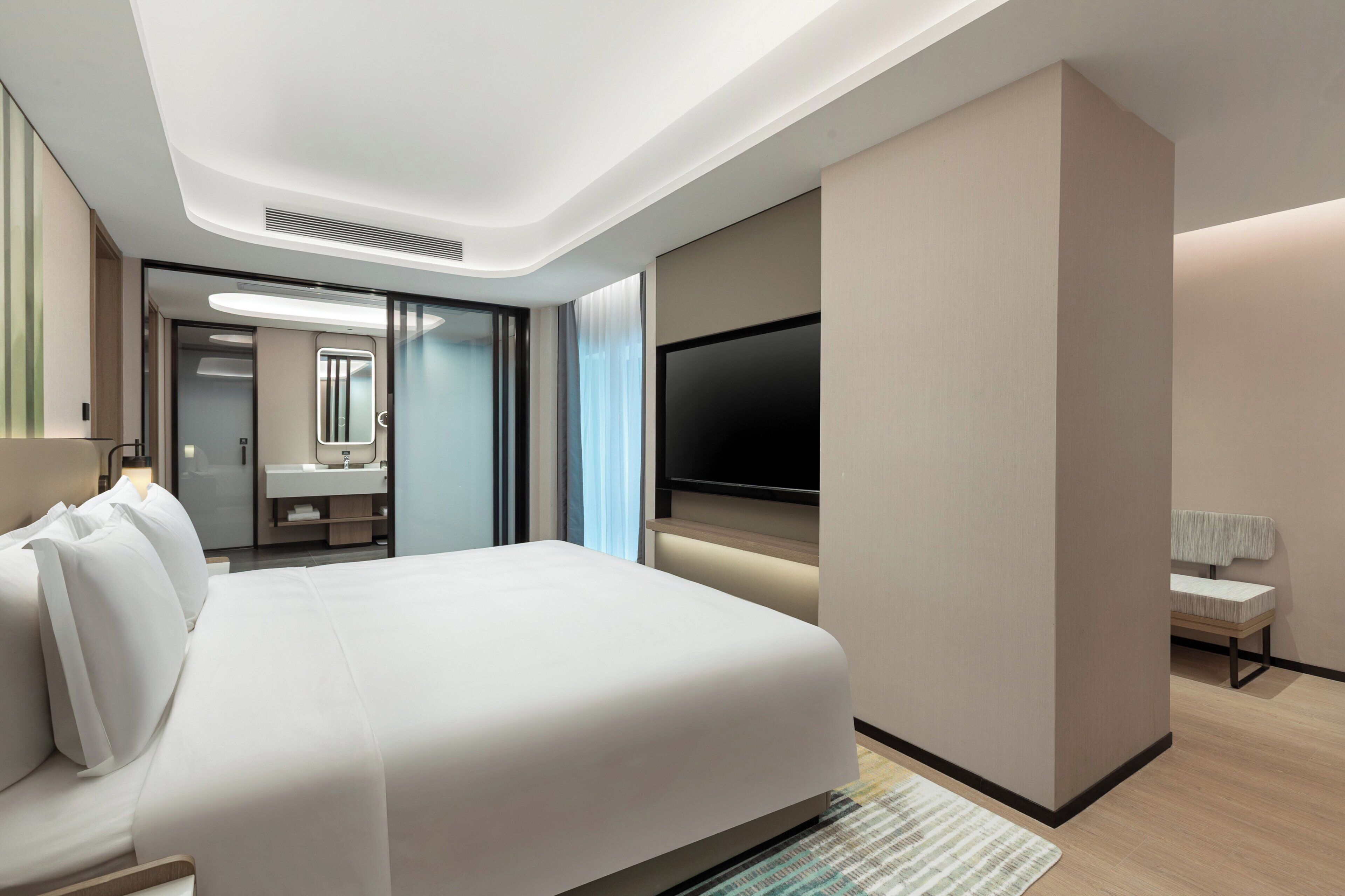 Habitación de hotel moderna con una cama grande, un televisor de pantalla plana en la pared, un baño con espejo y lavamanos, y una pequeña zona de estar con una mesa y una silla.