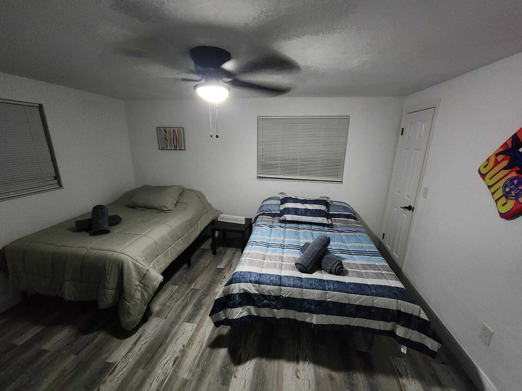 A small, dimly lit room with two single beds, a ceiling fan, and a window with blinds.