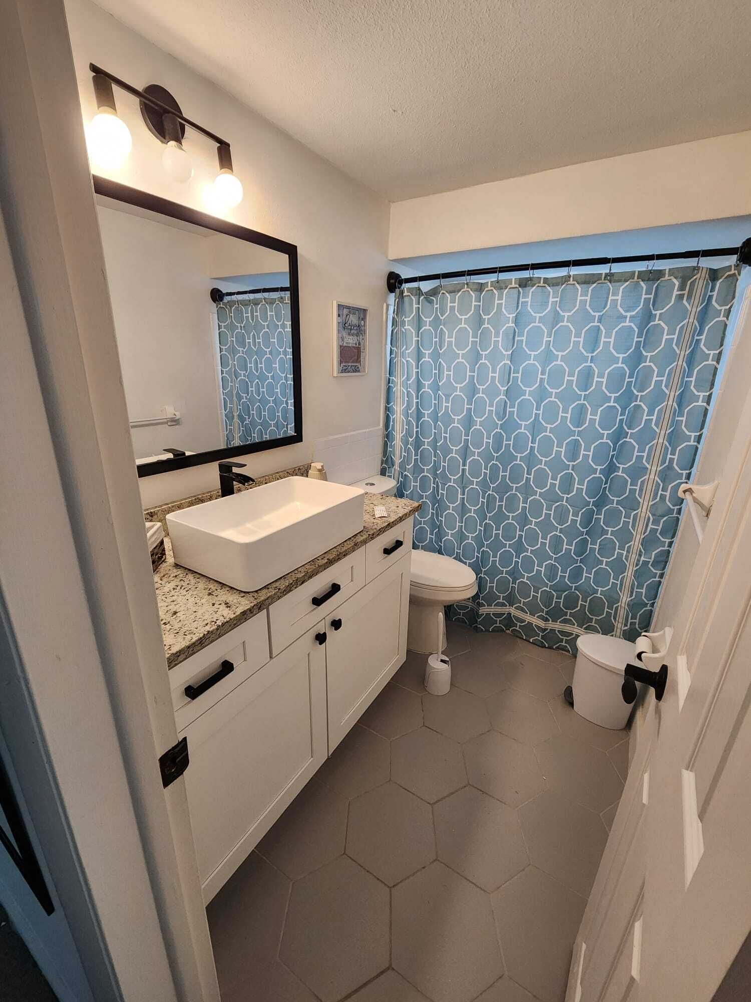 Une salle de bain avec un lavabo blanc, un miroir et des toilettes. Il y a un rideau de douche bleu orné d’un motif géométrique.