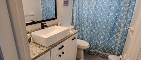 Une salle de bain avec un lavabo blanc, un miroir et des toilettes. Il y a un rideau de douche bleu orné d’un motif géométrique.