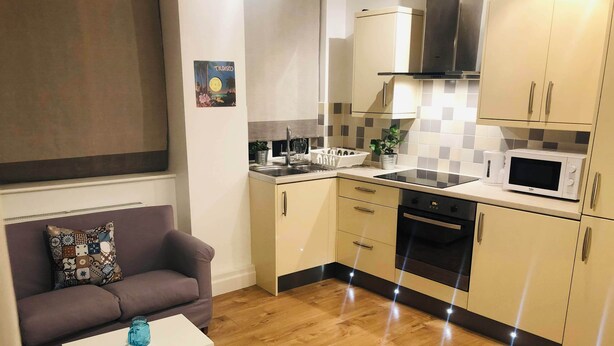 A compact kitchen with a microwave, oven, and sink. There's a sofa and a small table with a blue vase.
