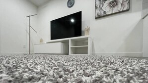 A room with a large flat-screen TV mounted on a white entertainment unit, a fluffy grey carpet, and a wall-mounted clock. - Comfy 2BR Basement Getaway (Edmonton)