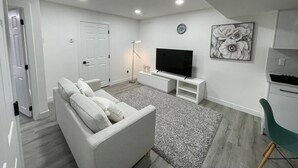 A modern living room with a white sofa, a flat-screen TV on a white stand, a floor lamp, and a painting on the wall. - Comfy 2BR Basement Getaway (Edmonton)