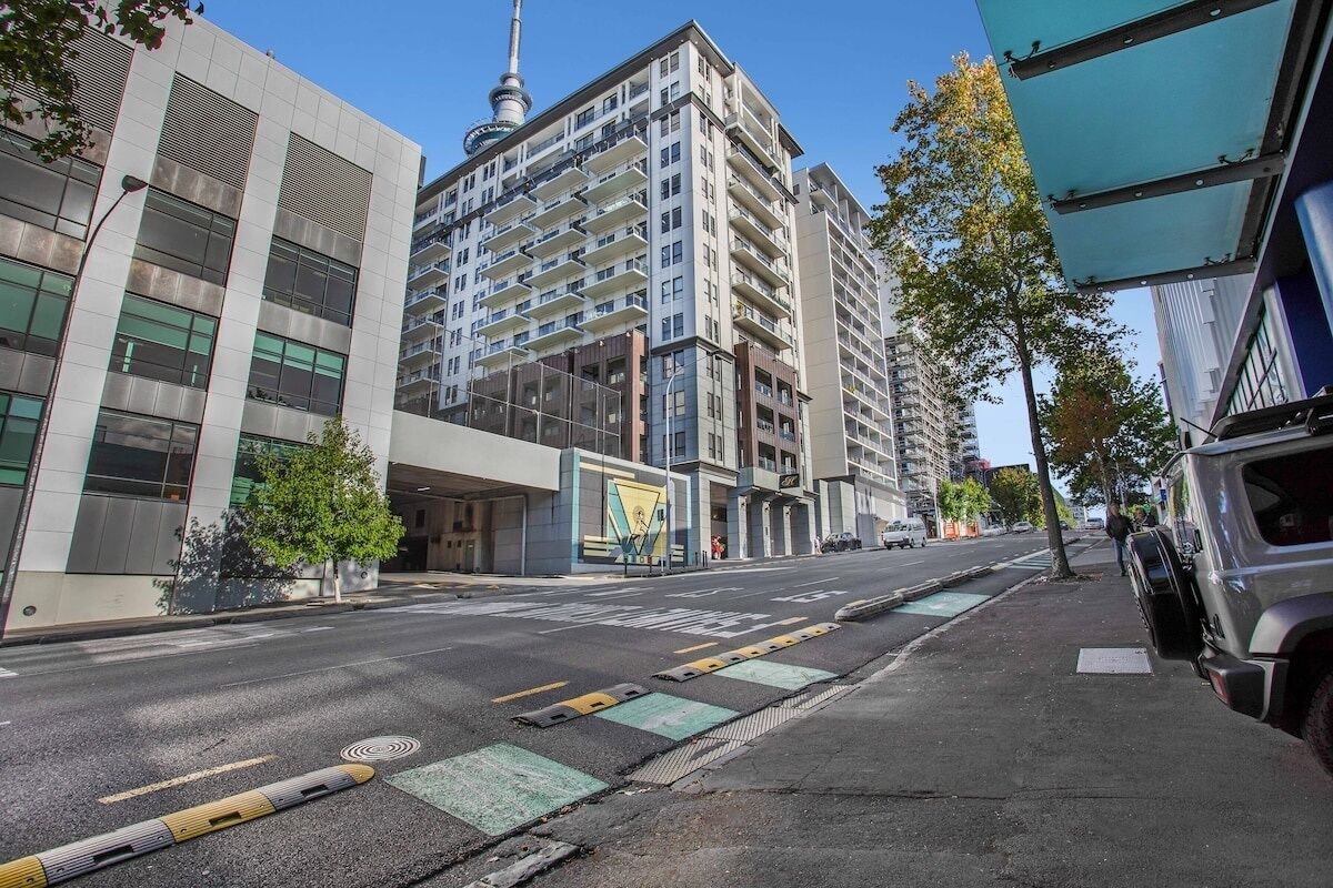 Uma rua urbana moderna com um edifício de vários andares, varandas e uma torre imponente.