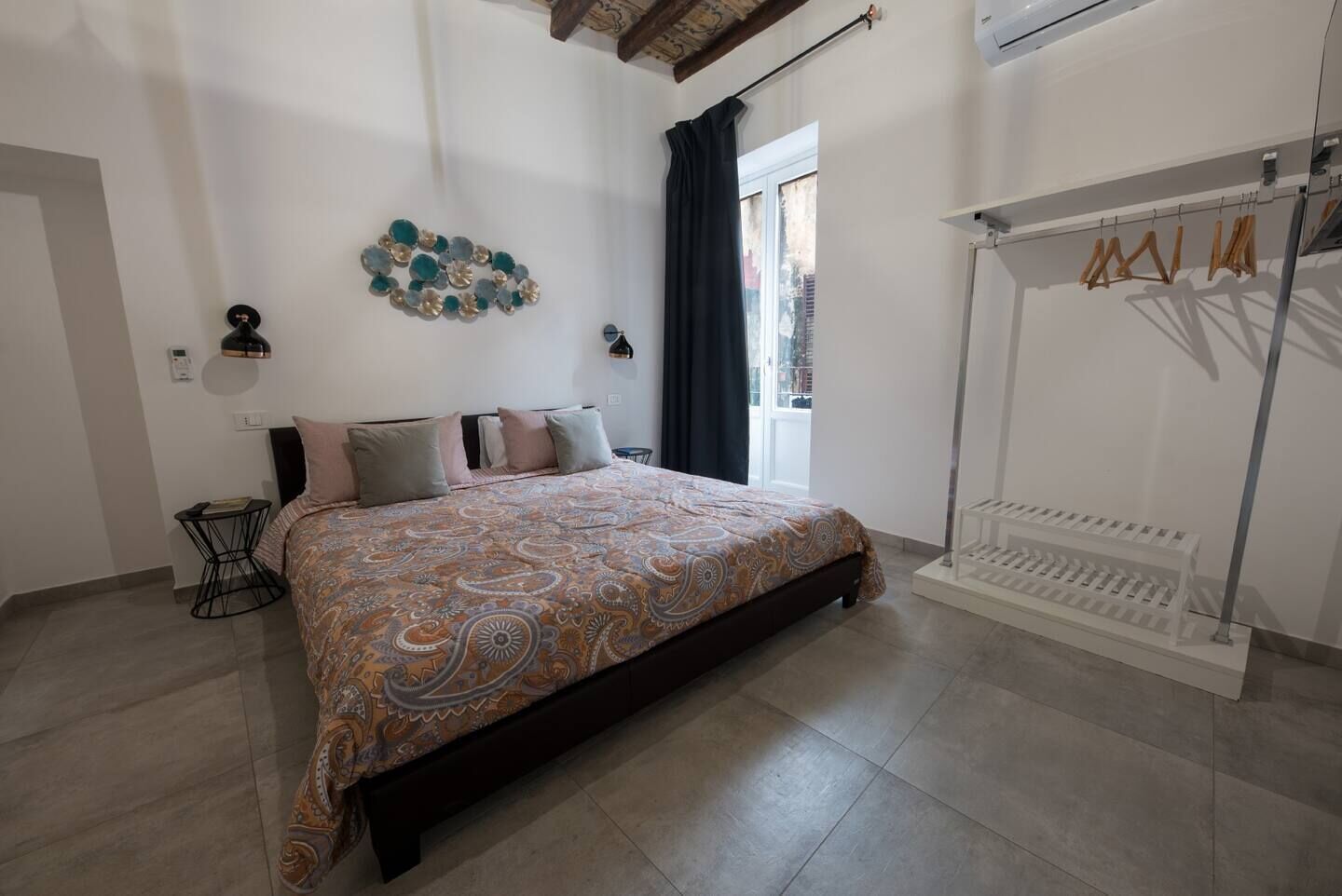 Une chambre à coucher comprenant un lit, une table de chevet, une lampe fixée au mur, une fenêtre avec des rideaux et un portant à vêtements.