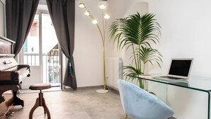 A modern room with a piano, a glass desk with a laptop, a blue chair, a tall floor lamp, and a large potted plant. - Casta Diva Luxury Apartments (Palermo)