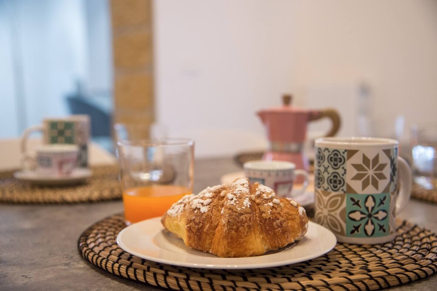 Un desayuno con un croissant en un plato, un vaso de zumo de naranja y tazas estampadas sobre un mantel tejido.