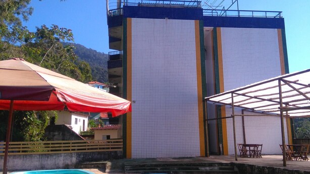 A pool area with lounge chairs, a red umbrella, and a building with a blue and white facade. - Baron Suítes (Ubatuba)