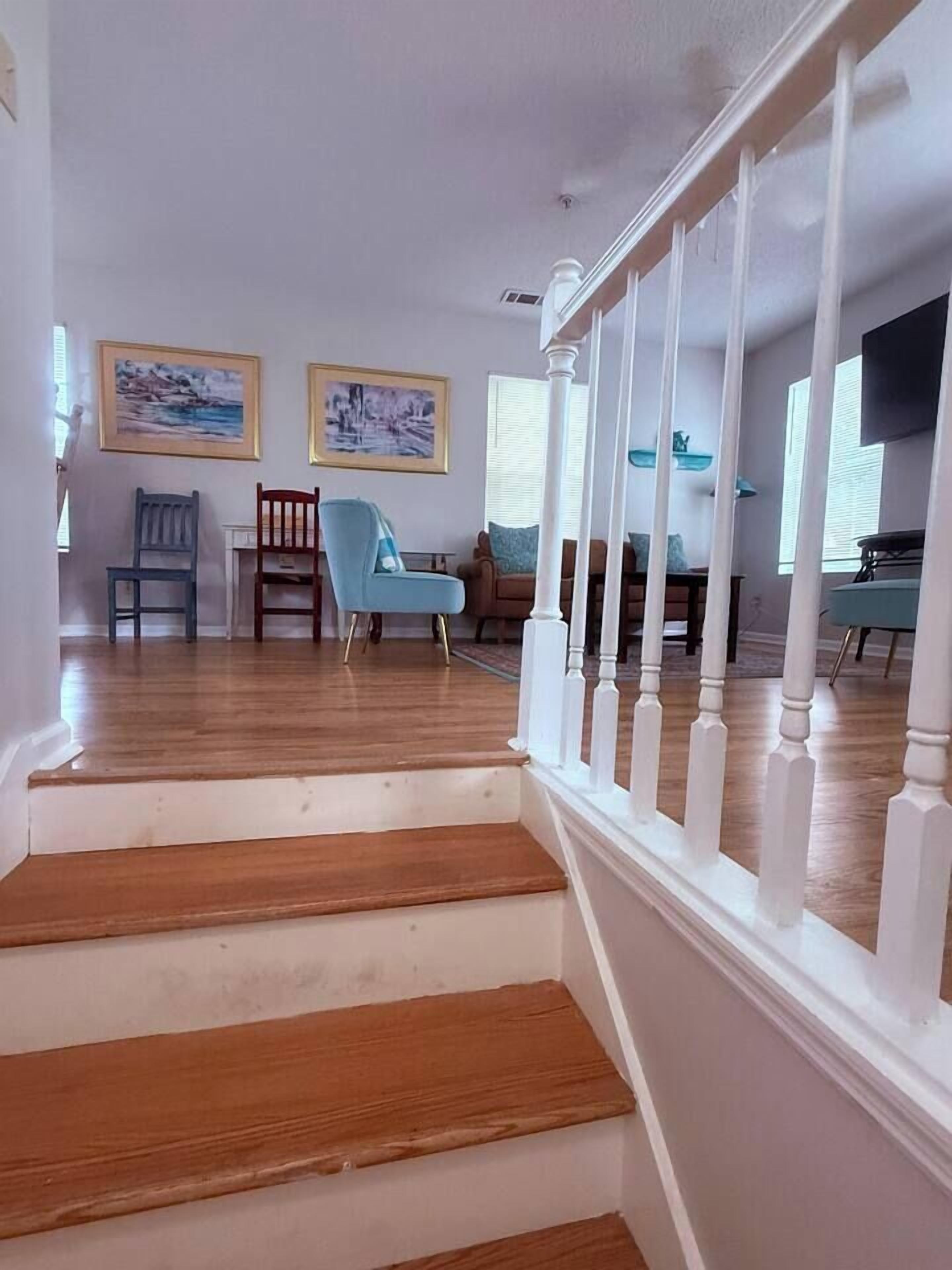 Un escalier avec des balustrades blanches mène à un espace de vie doté d’un plancher en bois, deux œuvres d’art encadrées ornent le mur et une chaise bleue s’y trouve.