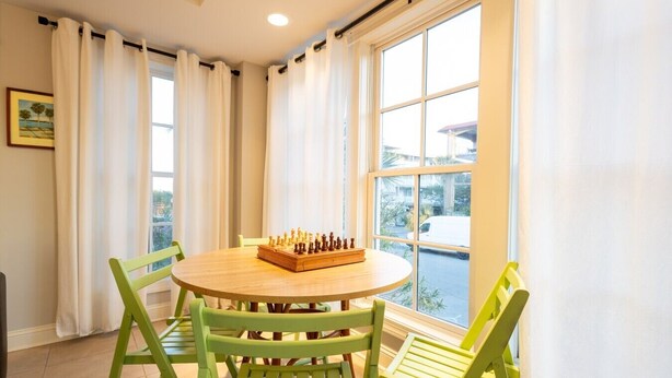 A room with a round wooden table, green chairs, a chessboard, and a large window with curtains.