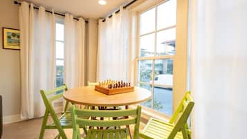 Une pièce avec une table ronde en bois, des chaises vertes, un jeu d’échecs et une grande fenêtre avec des rideaux.