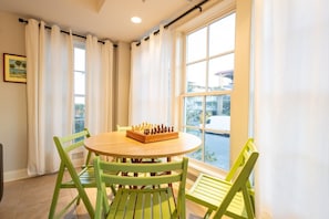 A room with a round wooden table, green chairs, a chessboard, and a large window with curtains.