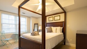 A four-poster bed with a headboard, a window with blinds, and a small table with chairs.