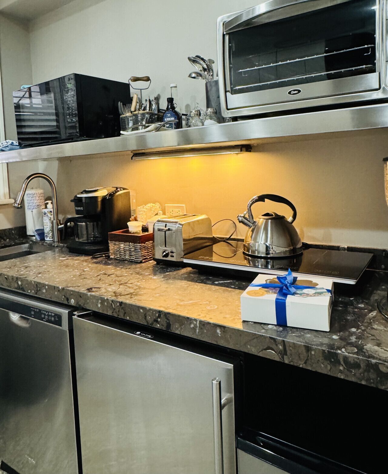 A kitchen with a microwave, toaster, kettle, and a dish rack. There is a box with a blue ribbon on the counter.