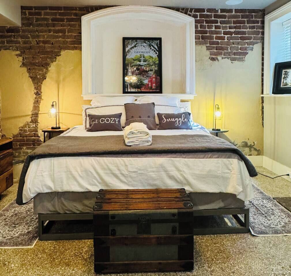 Una cama con cabecera, almohadas con texto, una toalla doblada y una mesita de noche con una lámpara.