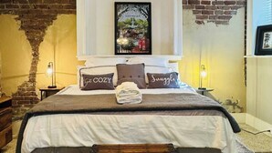 Uma cama com cabeceira, travesseiros com texto, uma toalha dobrada e uma mesa de cabeceira com uma luminária.