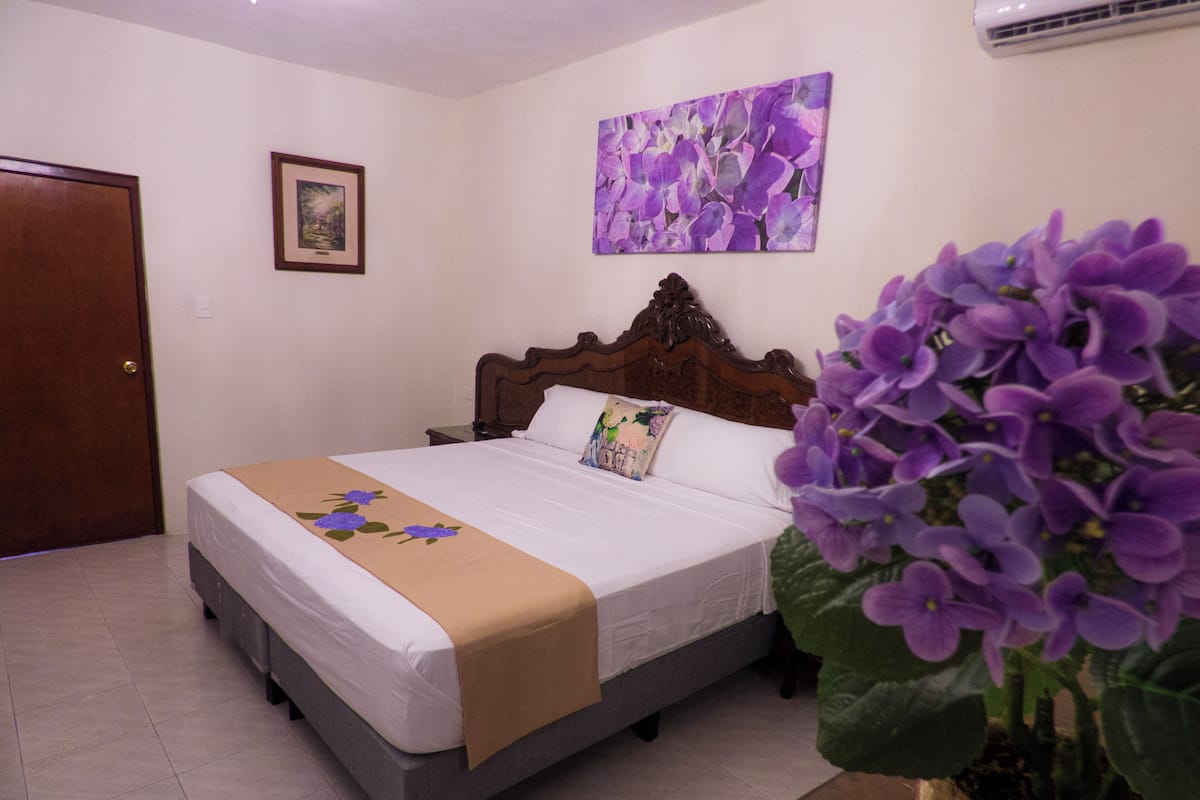 Une chambre à coucher avec un lit, une tête de lit en bois, un tableau encadré accroché au mur et une plante en pot ornée de fleurs violettes.