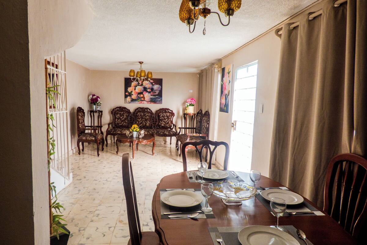 Une salle à manger avec une table ronde dressée pour six personnes, entourée de chaises en bois. Un lustre est suspendu au-dessus de la table, et un tableau est accroché au mur derrière l’espace où sont placées les chaises.