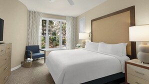 A hotel room with a large bed, a ceiling fan, a window with curtains, a desk with a lamp, and a TV on a dresser.
