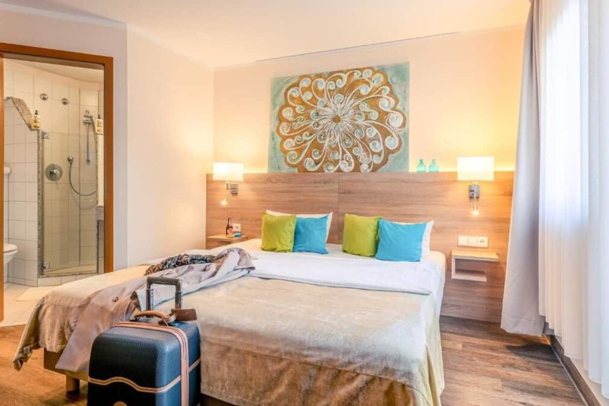 A hotel room with a bed, a shower area, a decorative wall piece, and a suitcase.