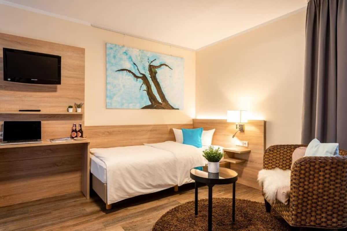 A hotel room with a bed, a desk with a laptop, a TV, a painting, a lamp, and a wicker chair.