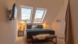 Ein Schlafzimmer mit Bett, zwei Dachfenstern, einer Bank, einem Fernseher und einem Kleiderständer.