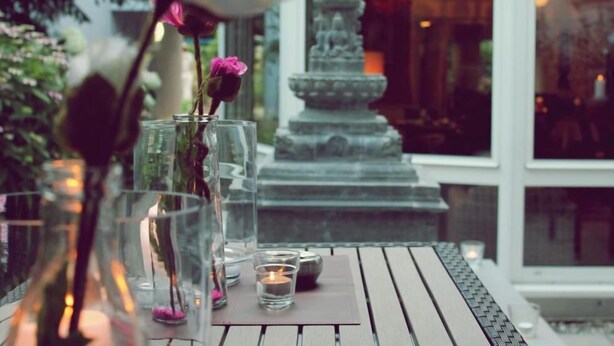 Ein Tisch mit Glasvasen, Kerzen, einer weißen Rose und einer Steinfontäne im Hintergrund.
