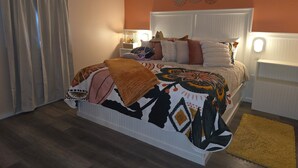 Un dormitorio con cama, mesitas de noche, un adorno en la pared y una ventana con cortinas.