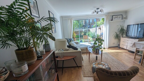 Una sala de estar con sofá, sillón y una planta grande en maceta. Hay un televisor y una estantería con libros y objetos decorativos.