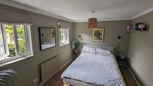 Un dormitorio con una cama, dos ventanas, un radiador, una lámpara colgante, una planta en maceta y una estantería.