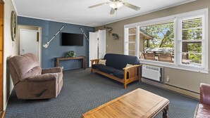 Un salon avec un ventilateur de plafond, un téléviseur fixé au mur, un canapé, un fauteuil et une table basse.
