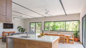 A modern kitchen with a central island, wooden cabinets, and large windows offering a view of greenery.