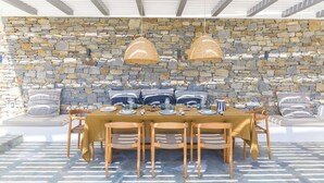 An outdoor dining area with a stone wall backdrop, wooden chairs, and a table set for a meal.