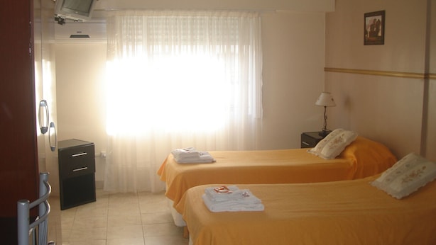 Une chambre d’hôtel avec deux lits, un téléviseur posé sur un meuble, un bureau avec une lampe et une fenêtre avec des rideaux.