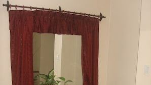 Une pièce avec un miroir, des rideaux rouges, une table à plateau de verre soutenue par deux colonnes de couleur dorée, et des plantes en pot.