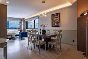 Un espace de repas moderne avec une table en bois, des chaises blanches, un canapé bleu et une vue sur la ville grâce à de grandes fenêtres.