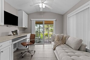 Un intérieur moderne avec un téléviseur à écran plat, un ordinateur portable, un bureau, un fauteuil de bureau marron, un canapé gris et un ventilateur de plafond.
