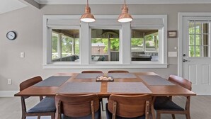A dining area with a wooden table, six chairs, two pendant lights, and large windows. - Serenity on Trinity - Hot Tub - Pets Welcome (Wilmington)