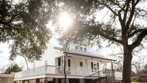 A white colonial-style house with a covered porch, a brick foundation, and a large tree in the foreground. - Little Noone by Raven’s Keep (Jackson)