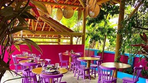 Un espace de restauration en plein air avec des chaises violettes et des tables rondes, entouré de verdure et agrémenté d’une pergola en bois.