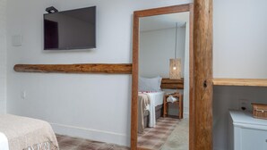 Une chambre avec une poutre en bois, un téléviseur à écran plat, un miroir et un lit doté d’une tête de lit blanche.