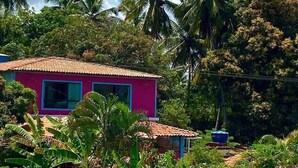 Une maison rose, avec un toit en tuiles, entourée d’une végétation luxuriante et de palmiers.