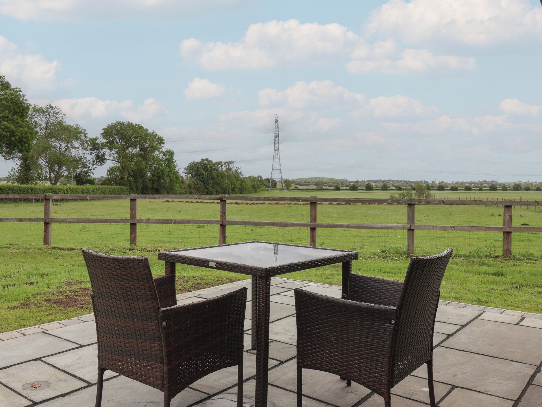 Een terras met twee rieten stoelen en een vierkante tafel, met uitzicht op een grasveld met een houten omheining en een elektriciteitspaal in de verte.
