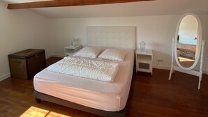 A bedroom with a bed, wooden floor, a dresser, a nightstand, a mirror, and a wooden beam ceiling.