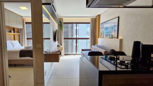 Habitación de hotel moderna con cama, sofá, zona de comedor con estufa y ventanas amplias con vistas a la ciudad.