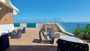 Terraza exterior iluminada por el sol con mobiliario de mimbre, barandilla de vidrio y vista al océano.