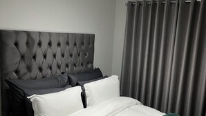 Une chambre à coucher avec une tête de lit capitonnée, un lit avec une literie blanche et verte, des rideaux gris et un luminaire au plafond.